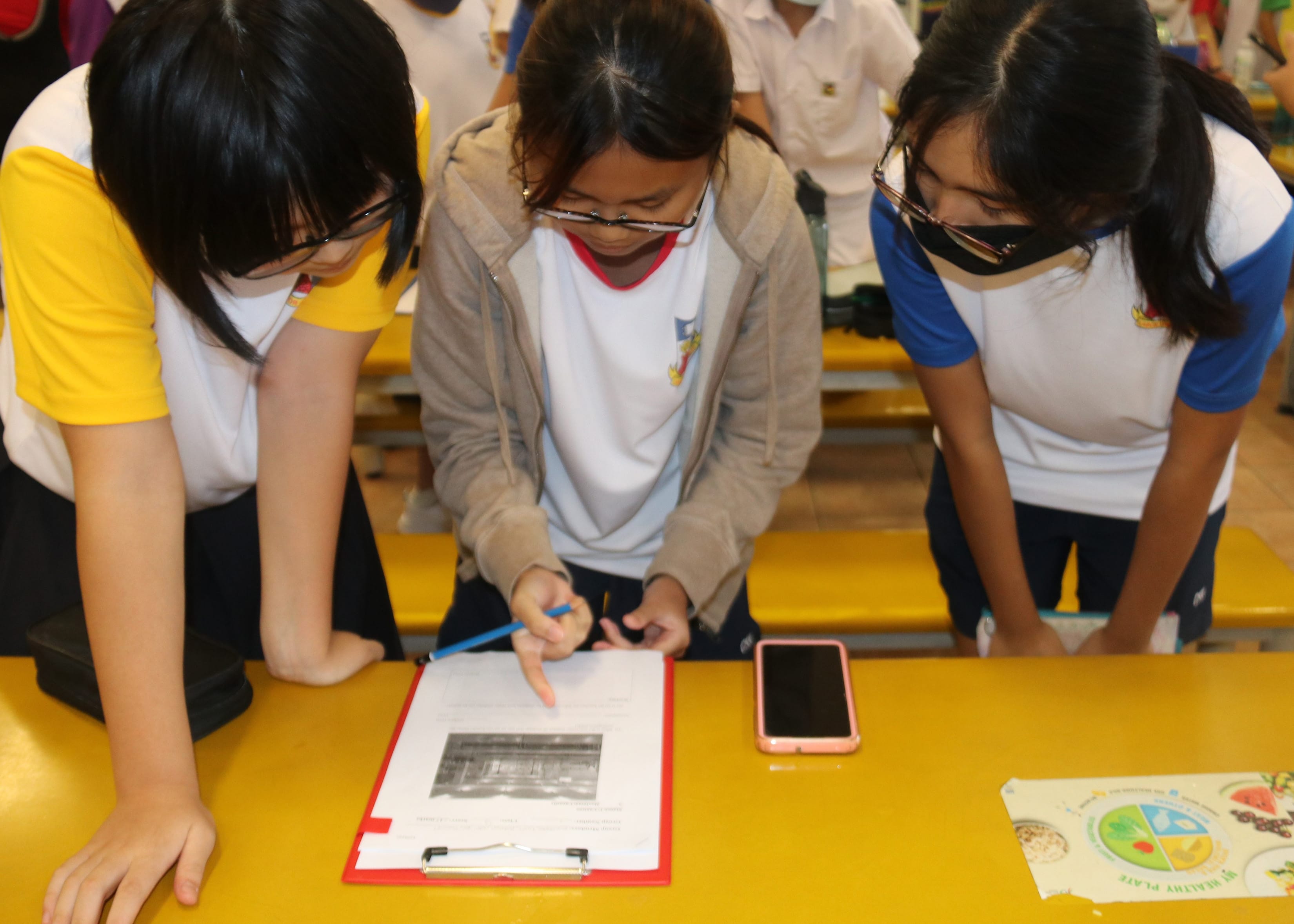 Three school girls lean over a yellow table, with one writing on a clipboard while others observe.
